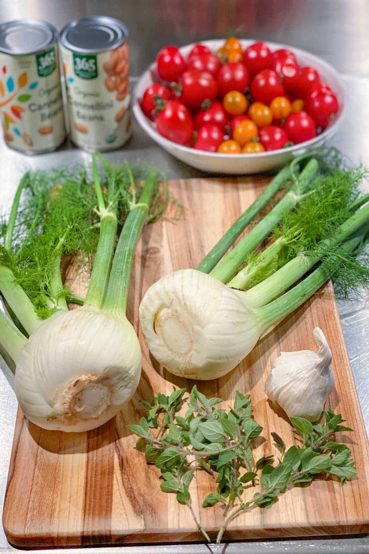 two whole fennel bulbs with fronds on a wooden cutting board with a whole bulb of garlic and several sprigs of fresh oregano, a bowl of cherry tomatoes and two cans of white beans in the background