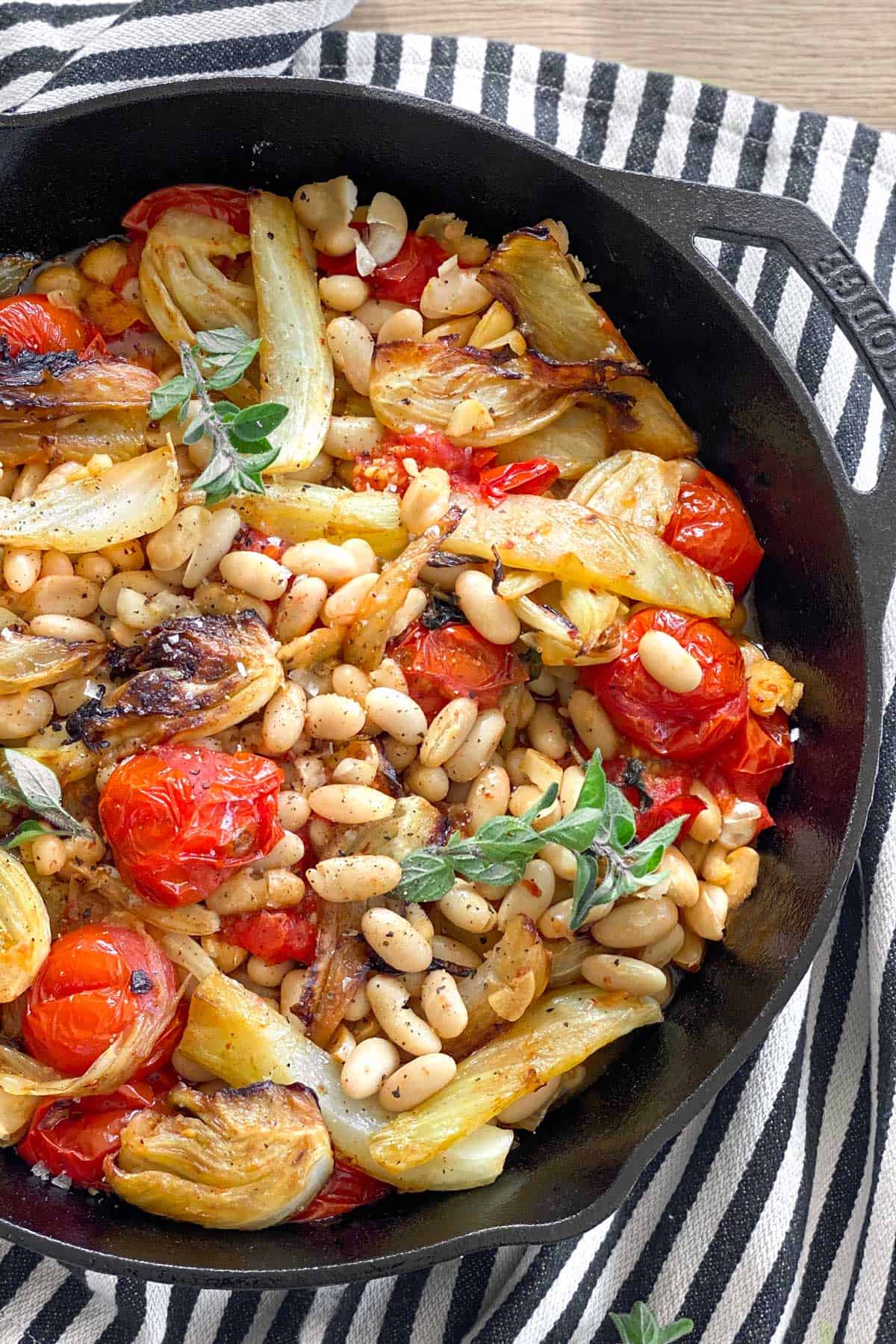 close up looking down into a cast iron skillet filled with roasted fennel, tomatoes and white beans, all set on a black and white striped dish towel.