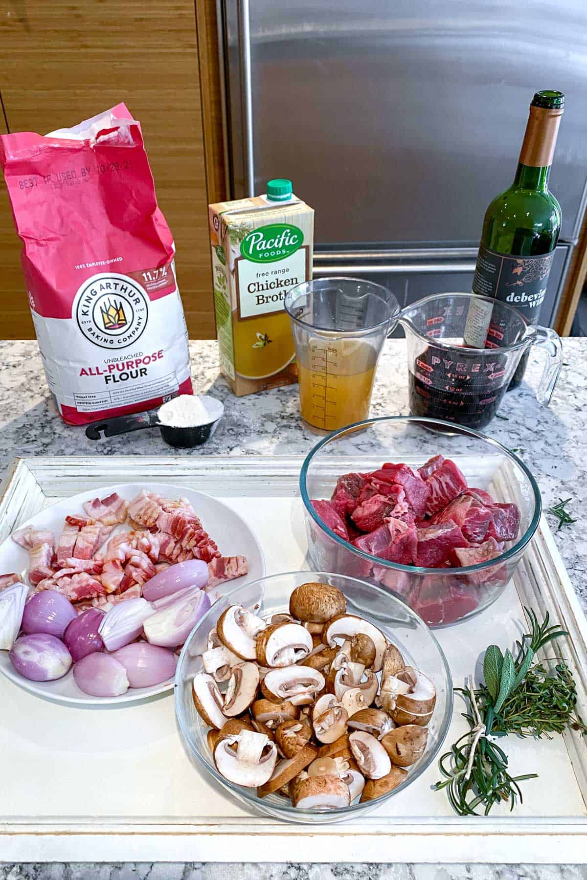 Ingredients for beef bourguignon on a white countertop: a bag of flour, glass bowl with cubed beef, bowl of sliced mushrooms, plate with chopped bacon and shallots, chicken broth in a clear cup, red wine in a clear cup, flour in a small black cup and herbs tied together