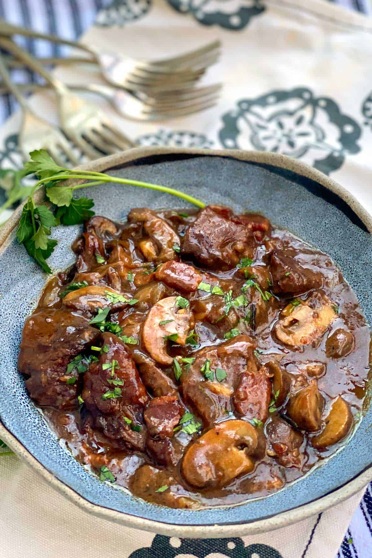 blue bowl filled with beef bourguignon, with a sprig of parsley on the side and a few forks in the background