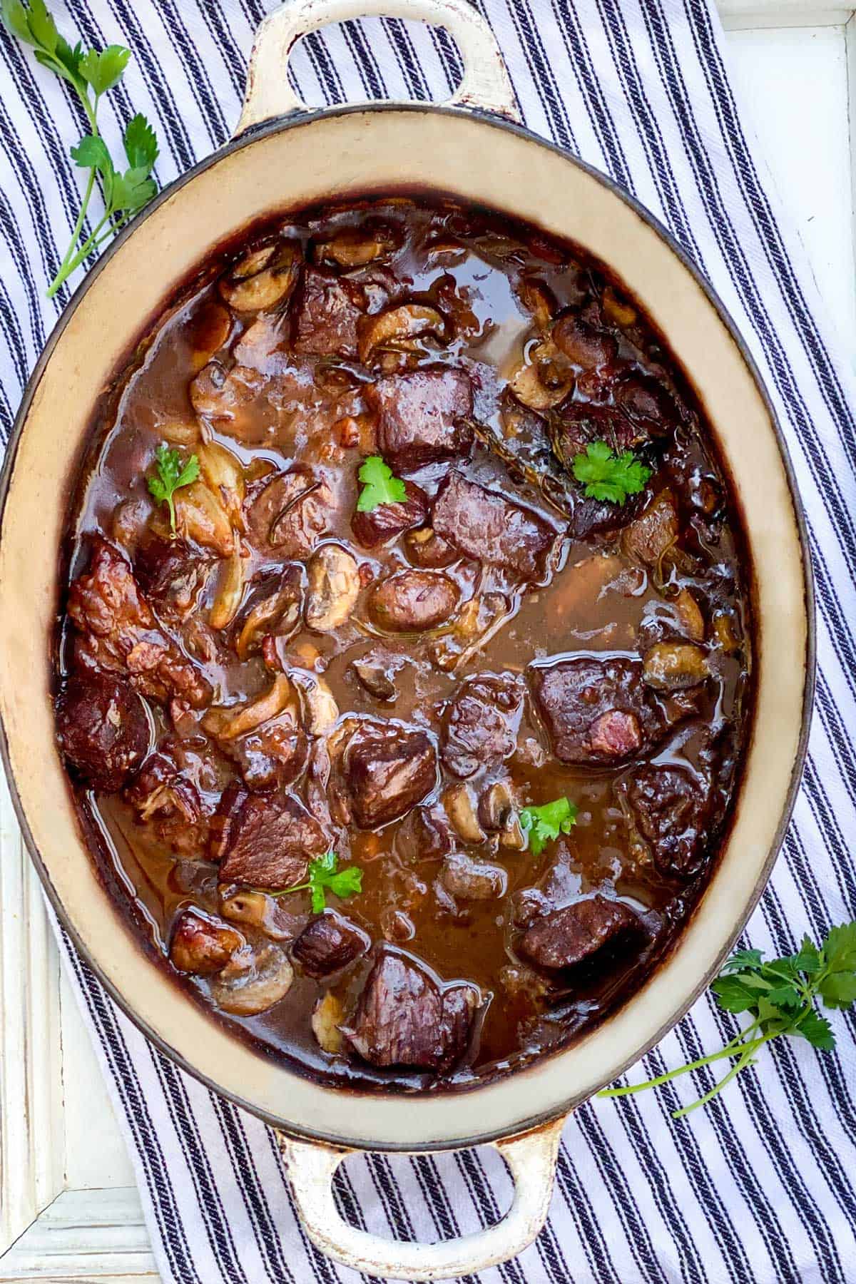 oval white Dutch oven on a black and white striped dish towel, seen from above, filled with beef bourguignon