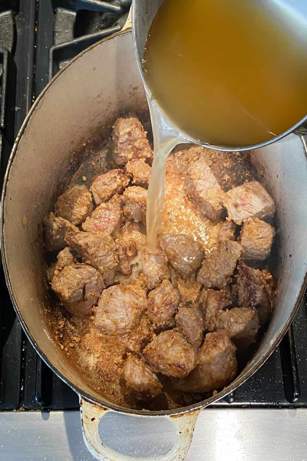hot chicken stock being poured into a stew pot over sautéed stew meat.