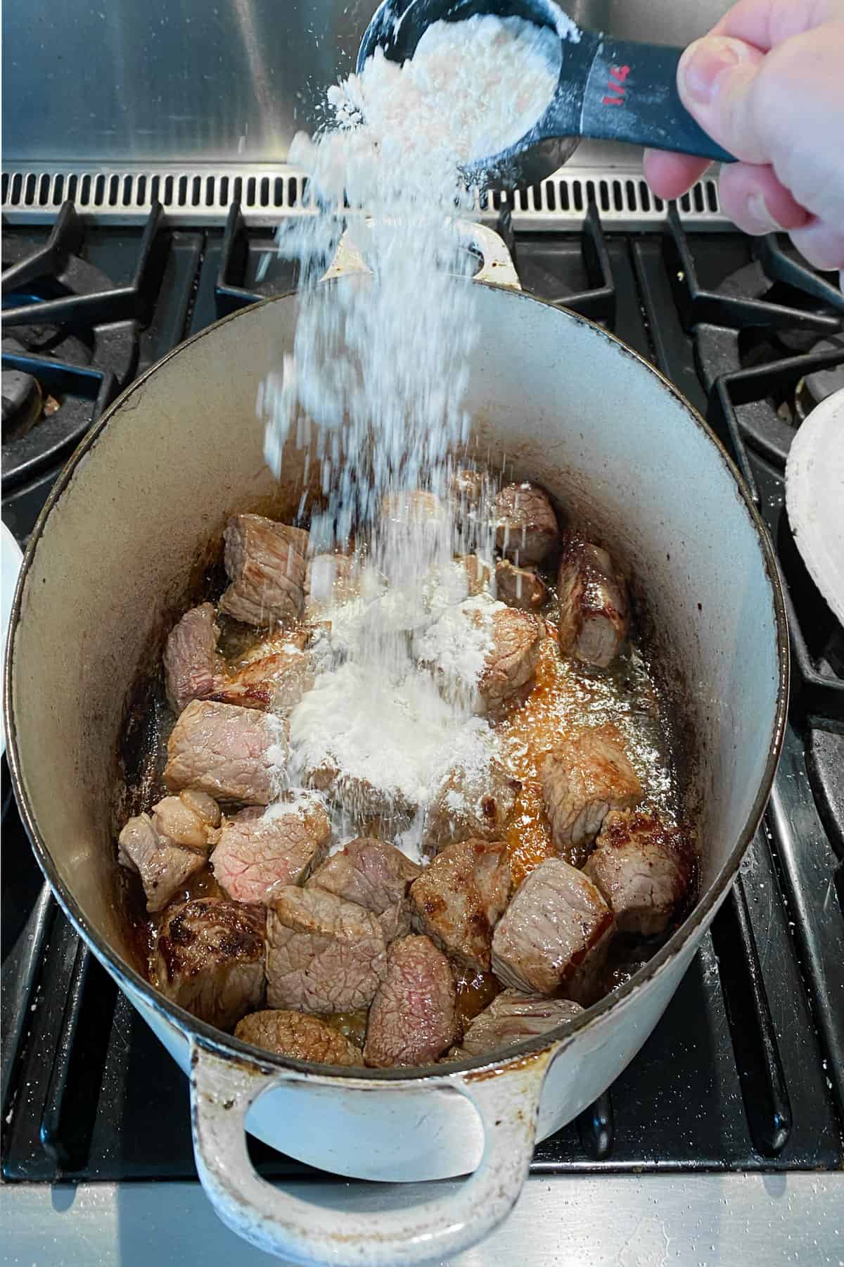 flour being sprinkled over browned beef cubes in a dutch oven.