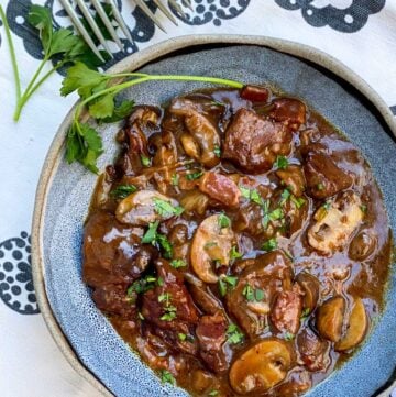 blue bowl filled with beef bourguignon, with a sprig of parsley on the side and a few forks in the background