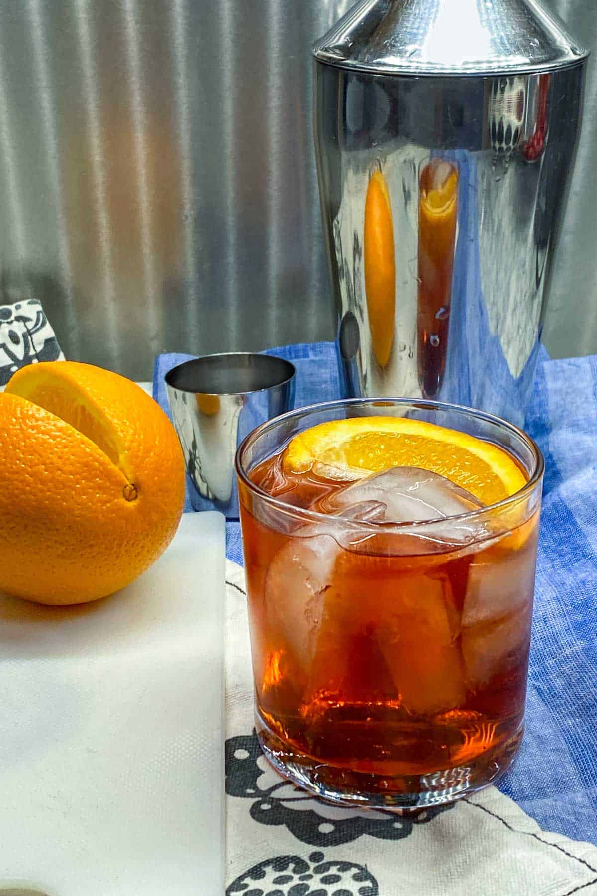 negroni cocktail in the foreground with an orange slice as a garnish, an orange and a cocktail shaker in the background