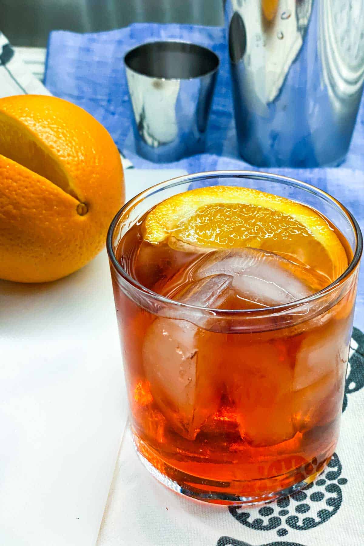 close up of a negroni cocktail in a glass with ice and an orange slice garnish, an orange and a cocktail shaker in the background, on a blue dish towel