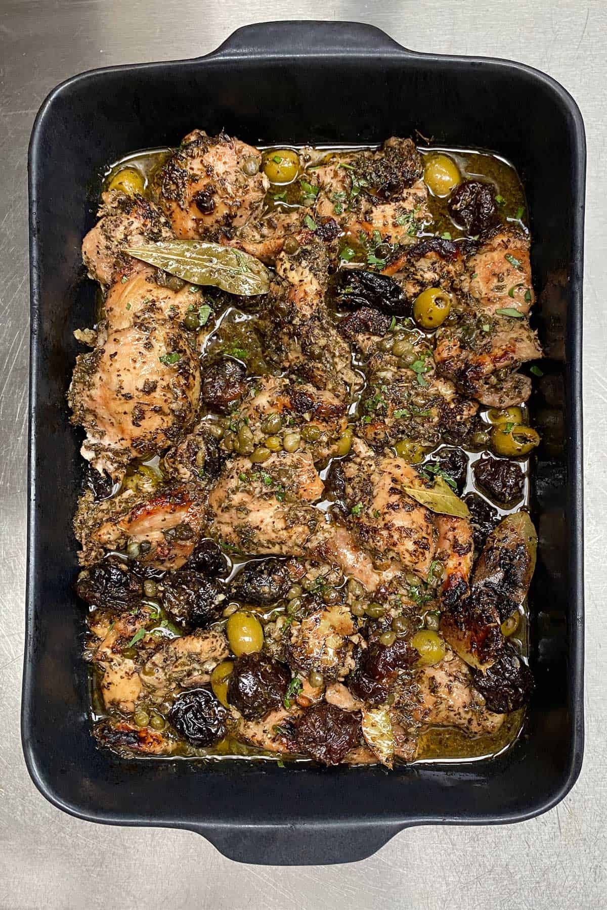 Rectangular black roasting pan filled with boneless chicken marbella, prunes, green olives and a bay leaf