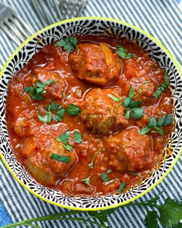 black and white decorative bowl filled with 6 Moroccan meatballs in tomato sauce, with chopped parsley sprinkled on top and a few forks in the background