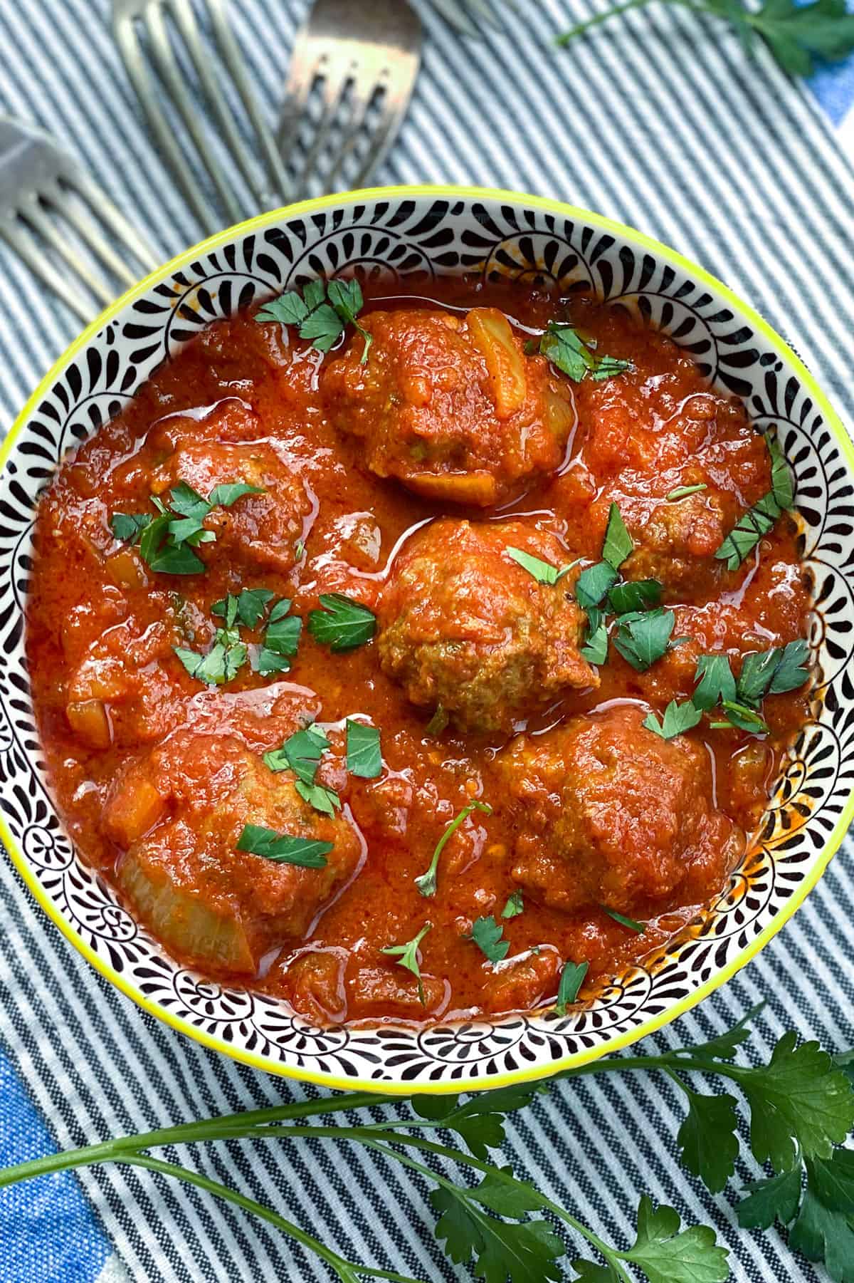 black and white decorative bowl filled with 6 Moroccan meatballs in tomato sauce, with chopped parsley sprinkled on top and a few forks in the background 