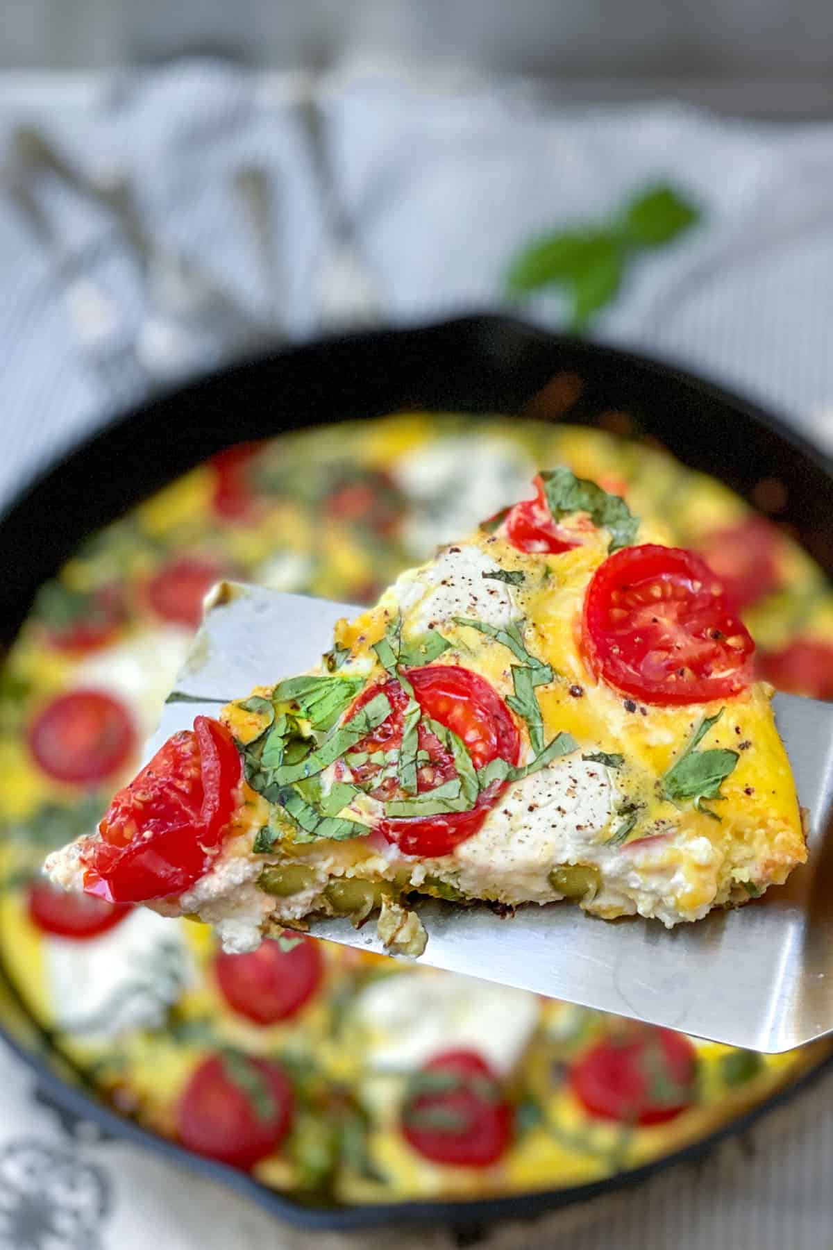 A slice of an asparagus goat cheese frittata with cherry tomatoes, on a spatula, held over the cast iron skillet containing the rest of the frittata