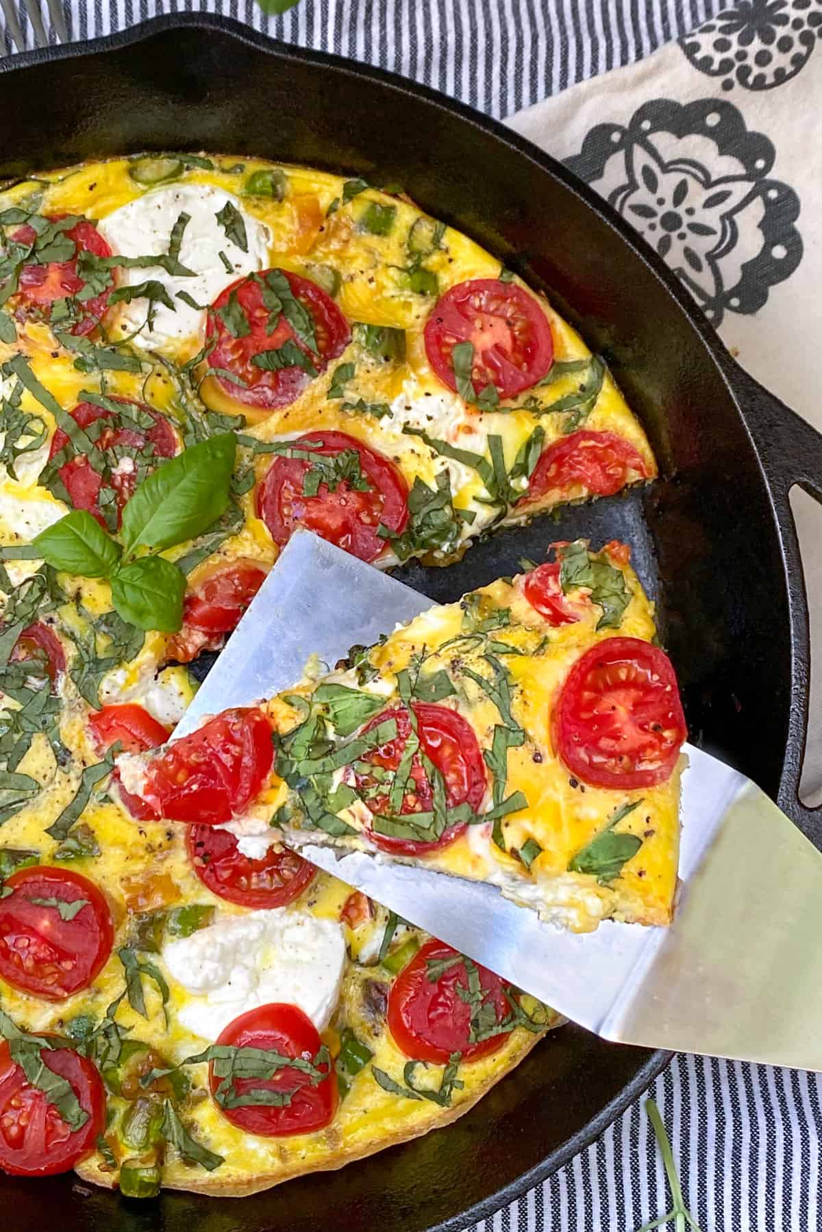 cast iron skillet filled with an asparagus frittata that's topped with cherry tomatoes and slivered basil, a spatula holds up one slice