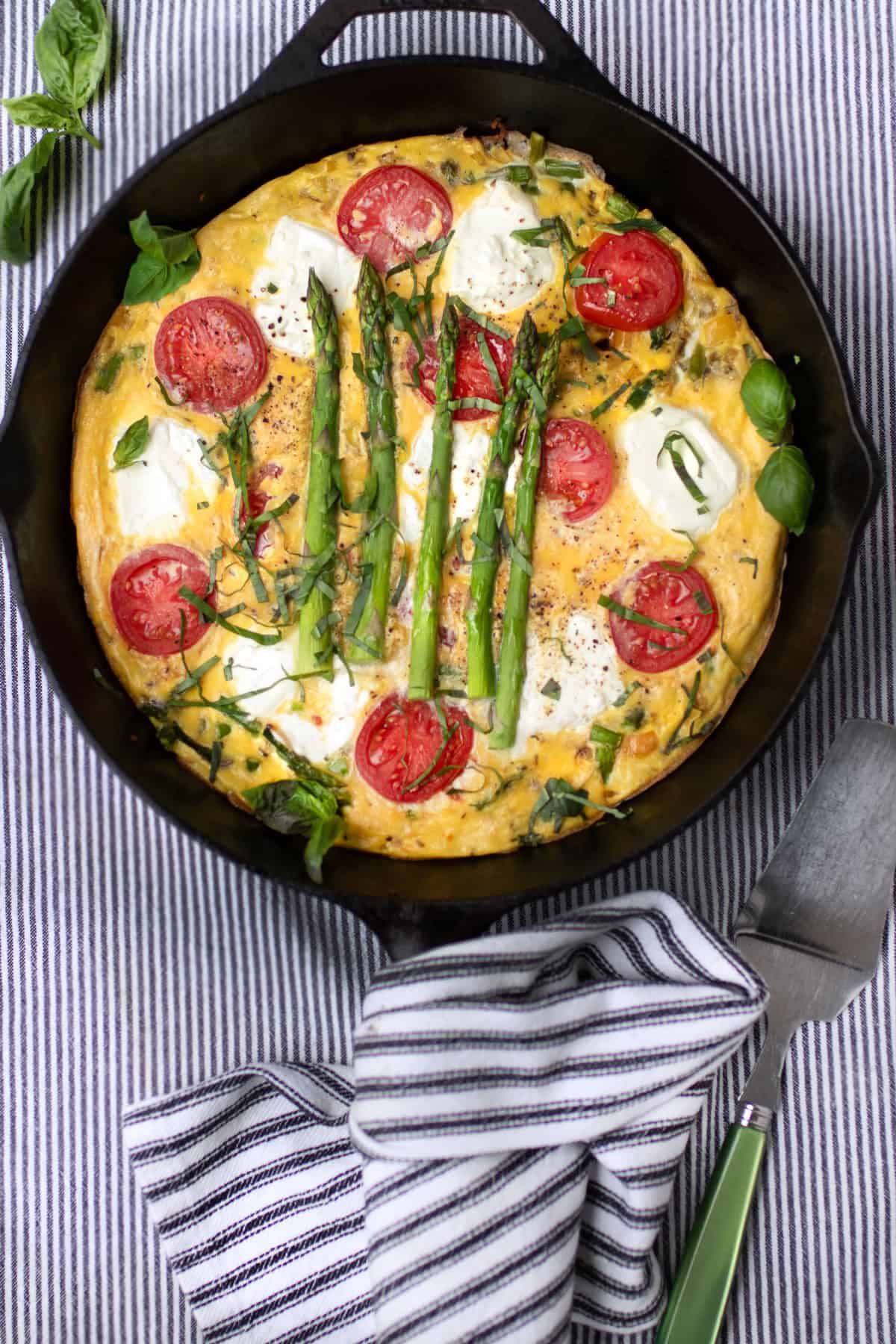 Cast iron skillet filled with a frittata decorated with rounds of goat cheese, sliced cherry tomatoes and 5 asparagus spears.