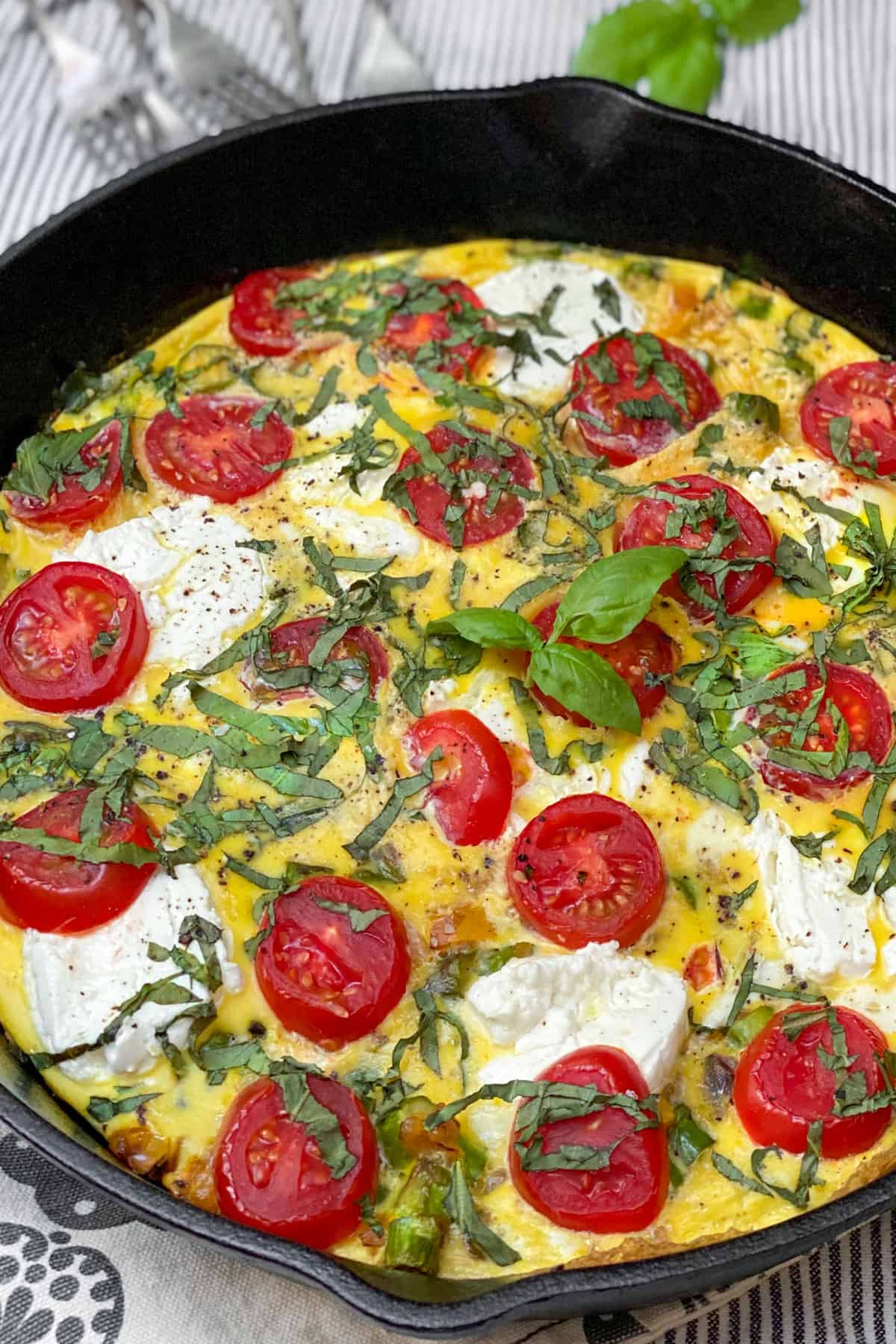 close up of an asparagus goat cheese frittata in a cast iron skillet topped with slices of cherry tomatoes and slivered basil.