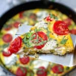 A slice of an asparagus goat cheese frittata with cherry tomatoes, on a spatula, held over the cast iron skillet containing the rest of the frittata