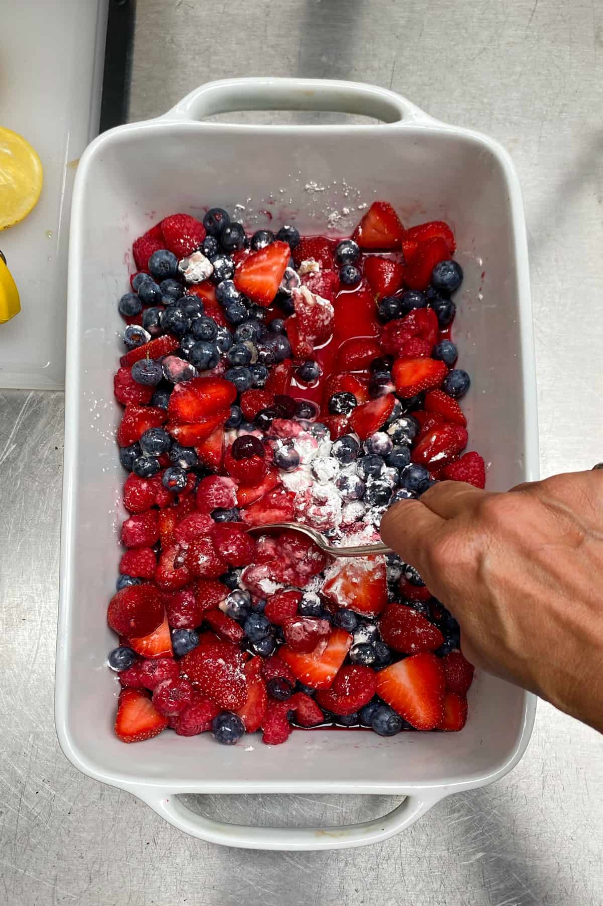 mixed berries - strawberries, raspberries and blueberries - in a rectangular porcelain baking pan. A hand is stirring in cornstarch and other dry ingredients.
