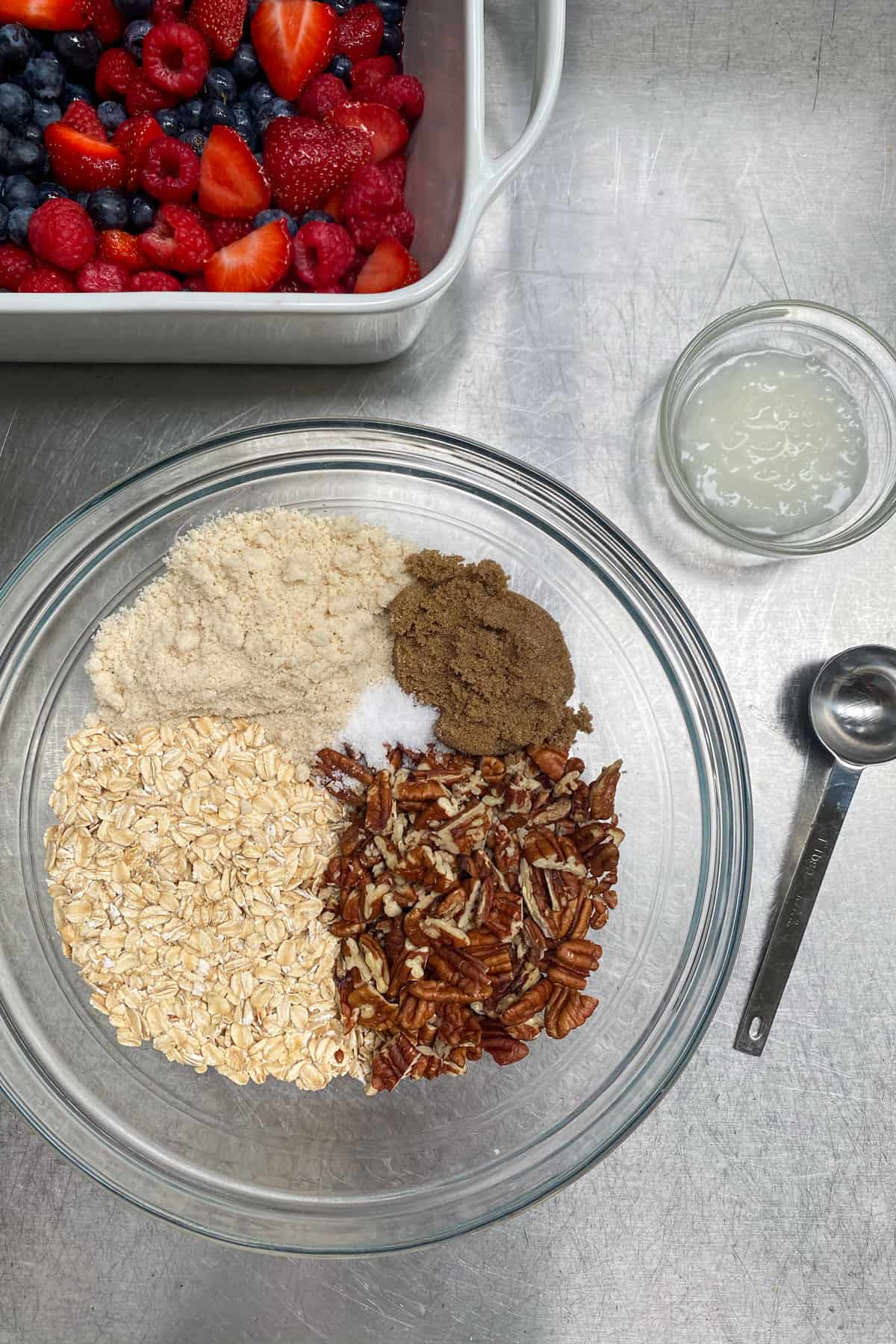 Topping ingredients for berry crisp in a glass bowl: chopped pecans, almond flour, oats, brown sugar and salt. A small bowl of melted coconut oil on the side.