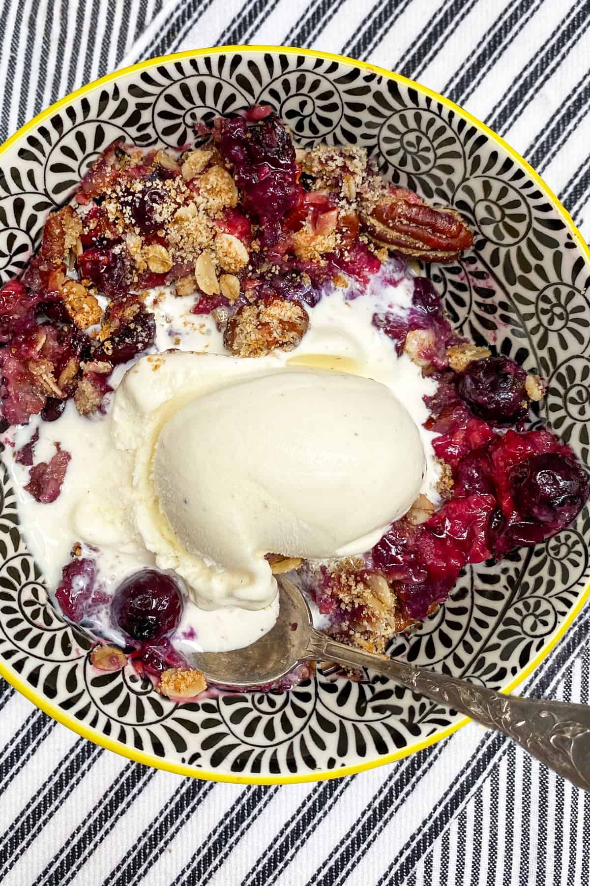 Mixed berry crumble in a bowl topped with ice cream, on a black and white striped dish cloth