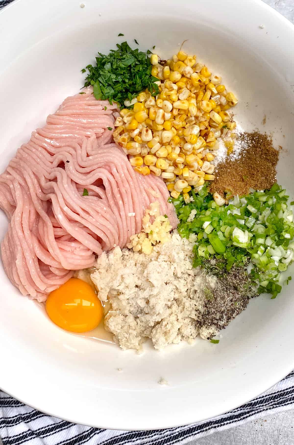 white mixing bowl with piles of ingredients: ground turkey, corn, an egg, chopped scallions, chopped parsley, soaked bread, spices