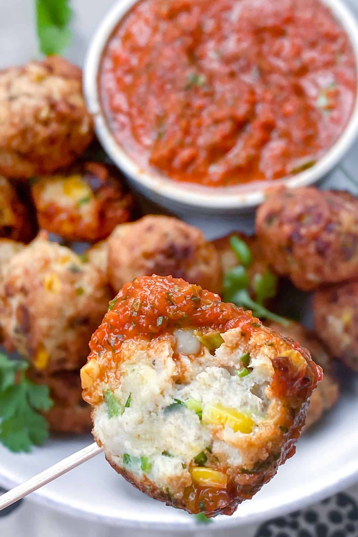 Close up of a turkey corn meatball with a bite take out of it, the platter of meatballs and red pepper sauce in the background