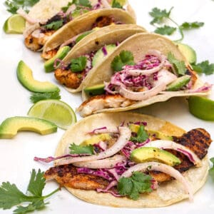5 blackened fish tacos lined up, filled with blackened fish, red cabbage slaw, slices of avocado, and cilantro leaves