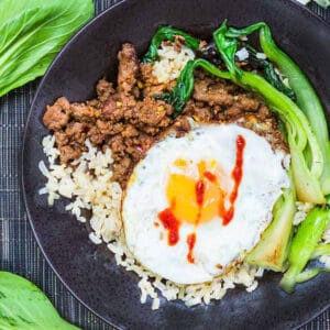 black plate topped with brown rice, sautéed ground beef, a fried egg with streaks of hot sauce and bok choy