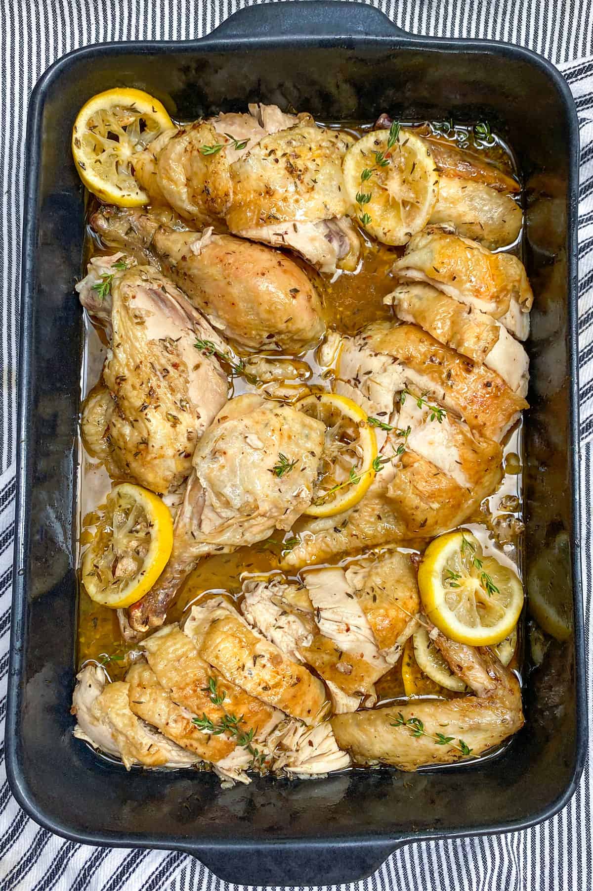 a whole slow roasted chicken cut into pieces in a black ceramic roasting pan with slice3s of lemon and sprigs of thyme strewn around.