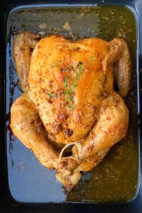 A whole roasted chicken in a black roasting pan with sprigs of thyme on top.