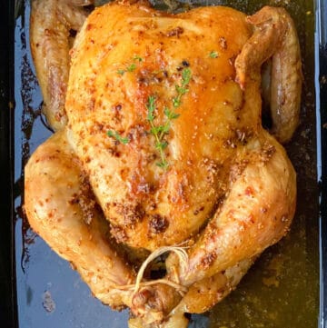 A whole roasted chicken in a black roasting pan with sprigs of thyme on top.