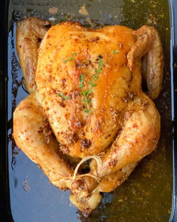 A whole roasted chicken in a black roasting pan with sprigs of thyme on top.