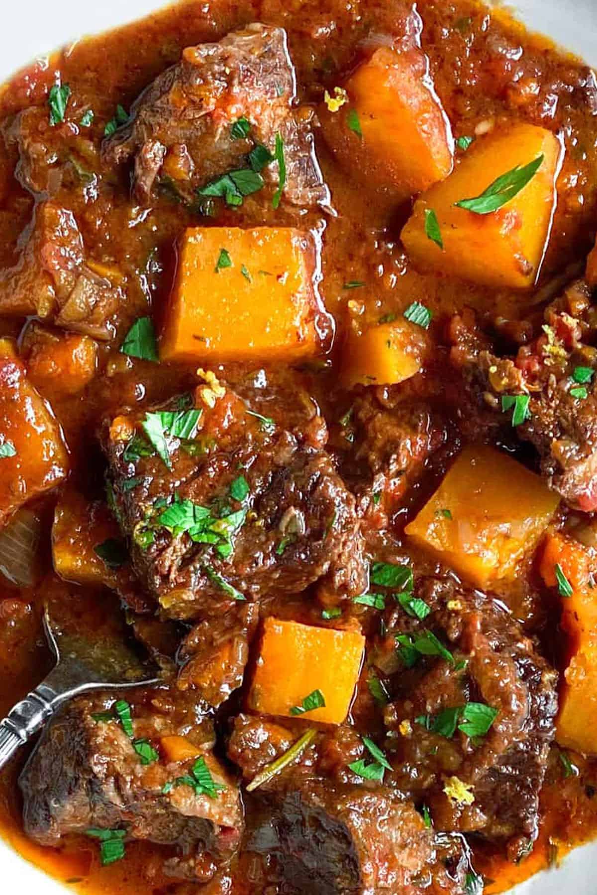 Close up of a bowl of stew with hunks of tender beef and cubes of butternut squash and a sprinkling of chopped parsley