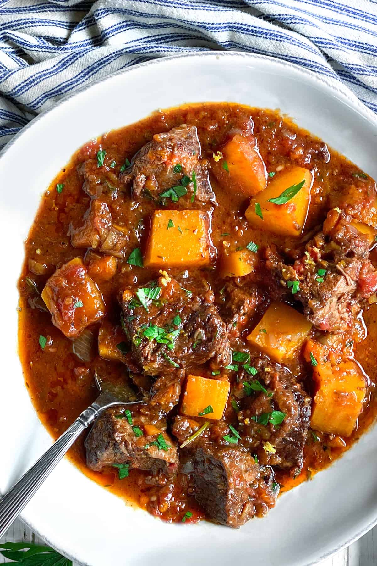 A white bowl of beef short ribs stew with butternut squash and a sprig of parsley