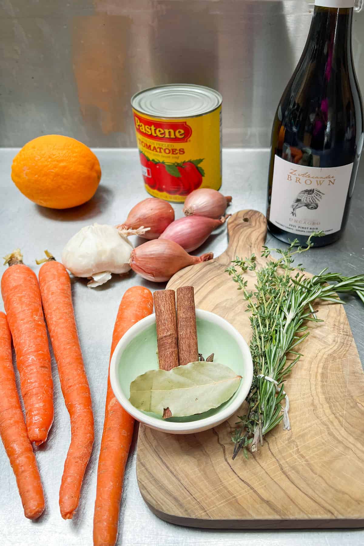 ingredients on a countertop: canned tomatoes, bottle of red wine, carrots, thyme, rosemary, an orange, shallots, garlic, cinnamon sticks, bay leaves and star anise