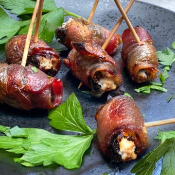 6 devils on horseback (bacon wrapped prunes) on a black plate with sprigs of parsley and toothpicks inserted into them
