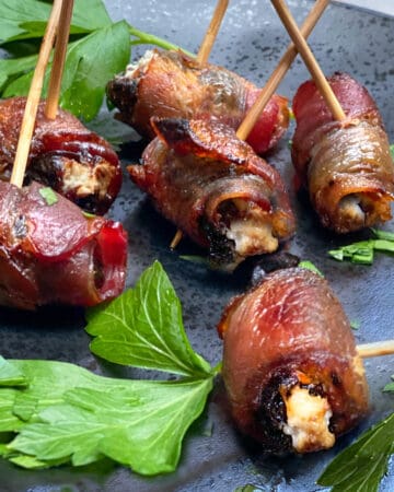 6 devils on horseback (bacon wrapped prunes) on a black plate with sprigs of parsley and toothpicks inserted into them