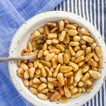 small white bowl on a blue dish towel, filled with toasted pine nuts