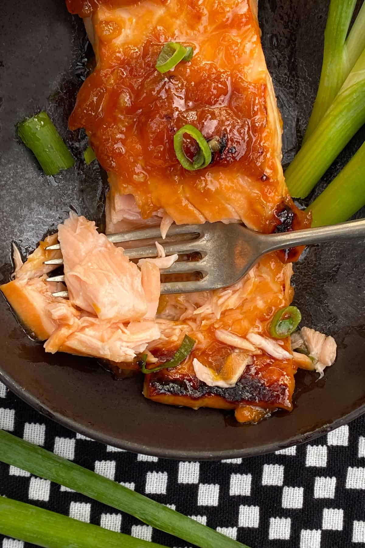 close up of a piece of apricot glazed broiled salmon in a black bowl, with a fork stuck through a tender looking piece of it