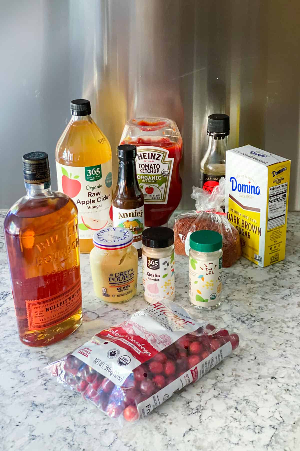 ingredients for cranberry bourbon bbq sauce sitting on a white countertop: a bottle of bourbon, bottle of ketchup, bag of fresh cranberries, bottle of cider vinegar, containers of garlic powder and onion powder, box of brown sugar.