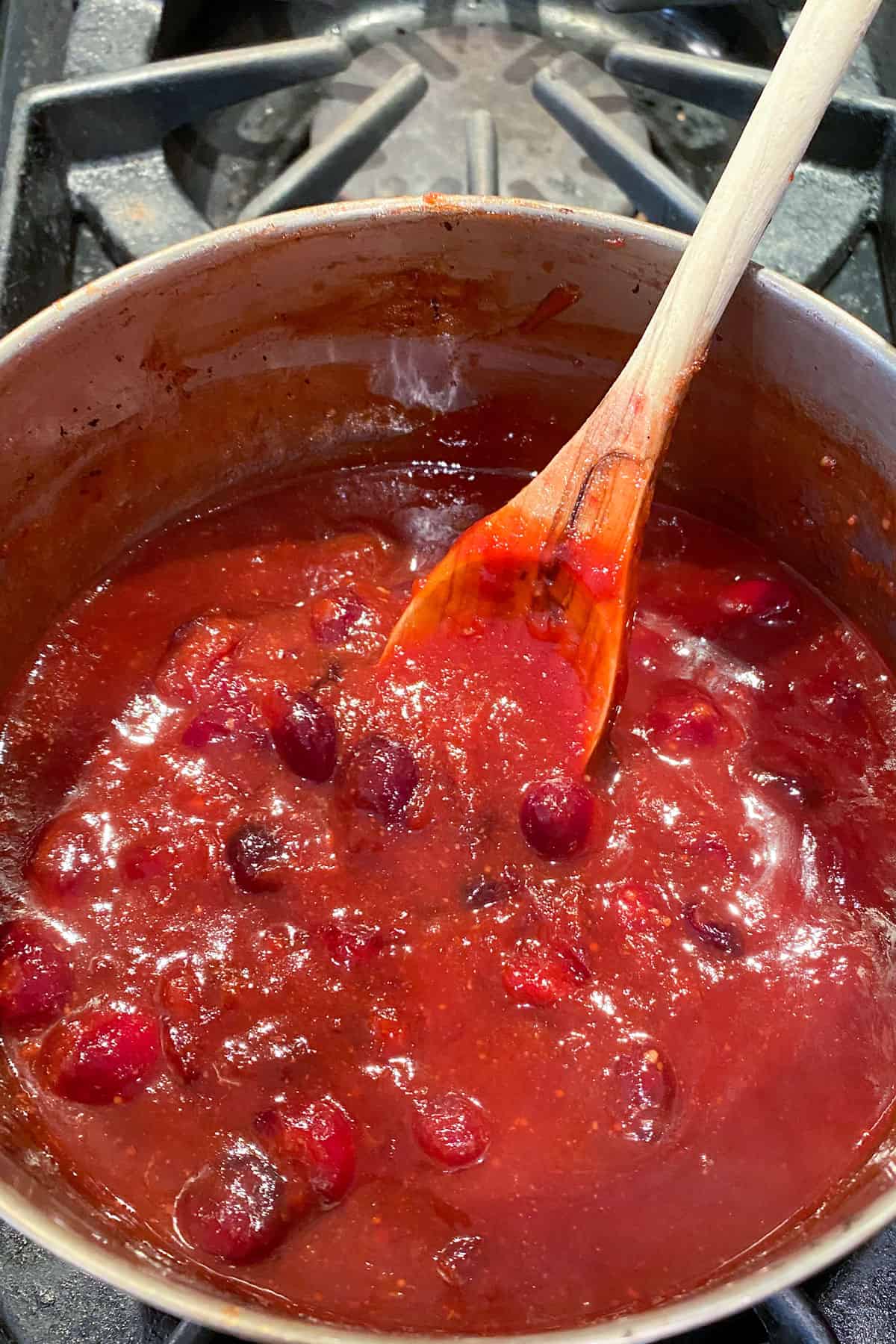 close up looking down into a saucepan filled with red cranberry bourbon bbq sauce, a wooden spoon is resting in the pot, the tip coated with the red sauce