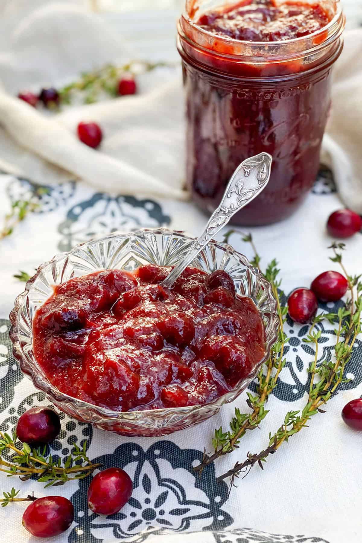 small glass bowl on a black and white cloth napkin, filled with cranberry bourbon bbq sauce, a few cranberries scattered around, a glass jar filled with cranberry sauce in the background.