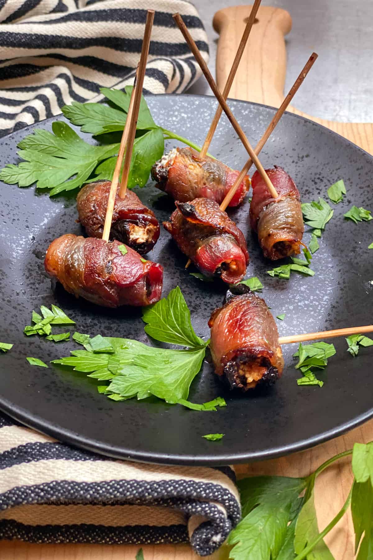 6 bacon wrapped prunes on a black plate with sprigs of parsley and toothpicks inserted into them