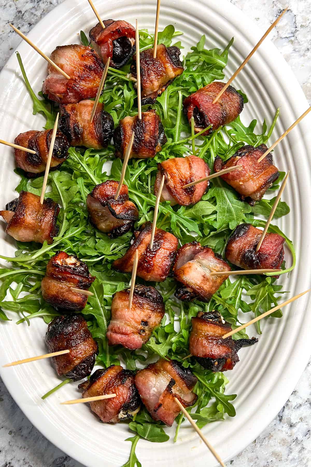 oval platter with about two dozen crispy bacon wrapped prunes on a bed of arugula, with toothpicks stuck into the bacon.