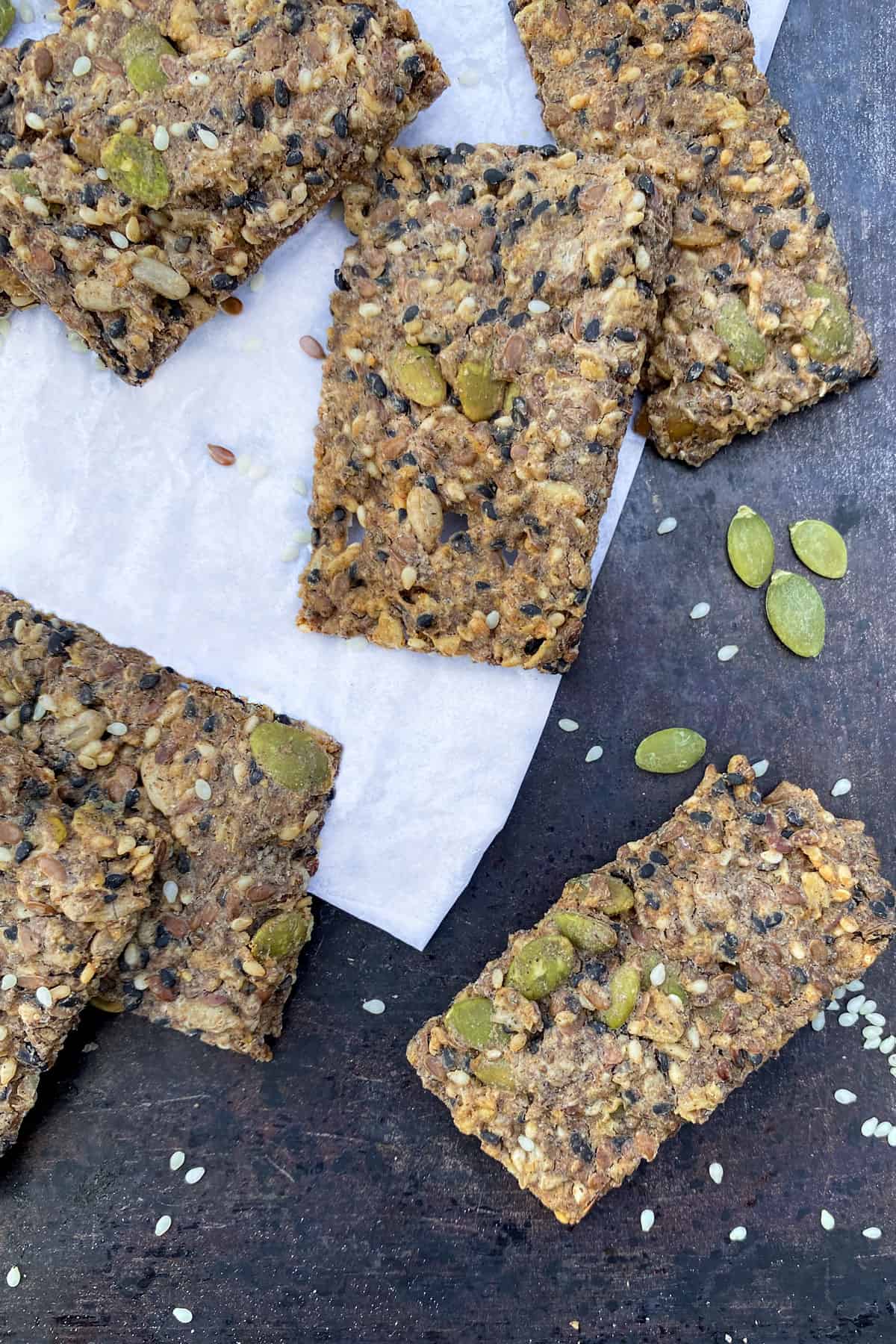 close up of 6 rectangular seeded crackers on a piece of white parchment, on a dark baking pan with a few pumpkin seeds and sesame seeds strewn around