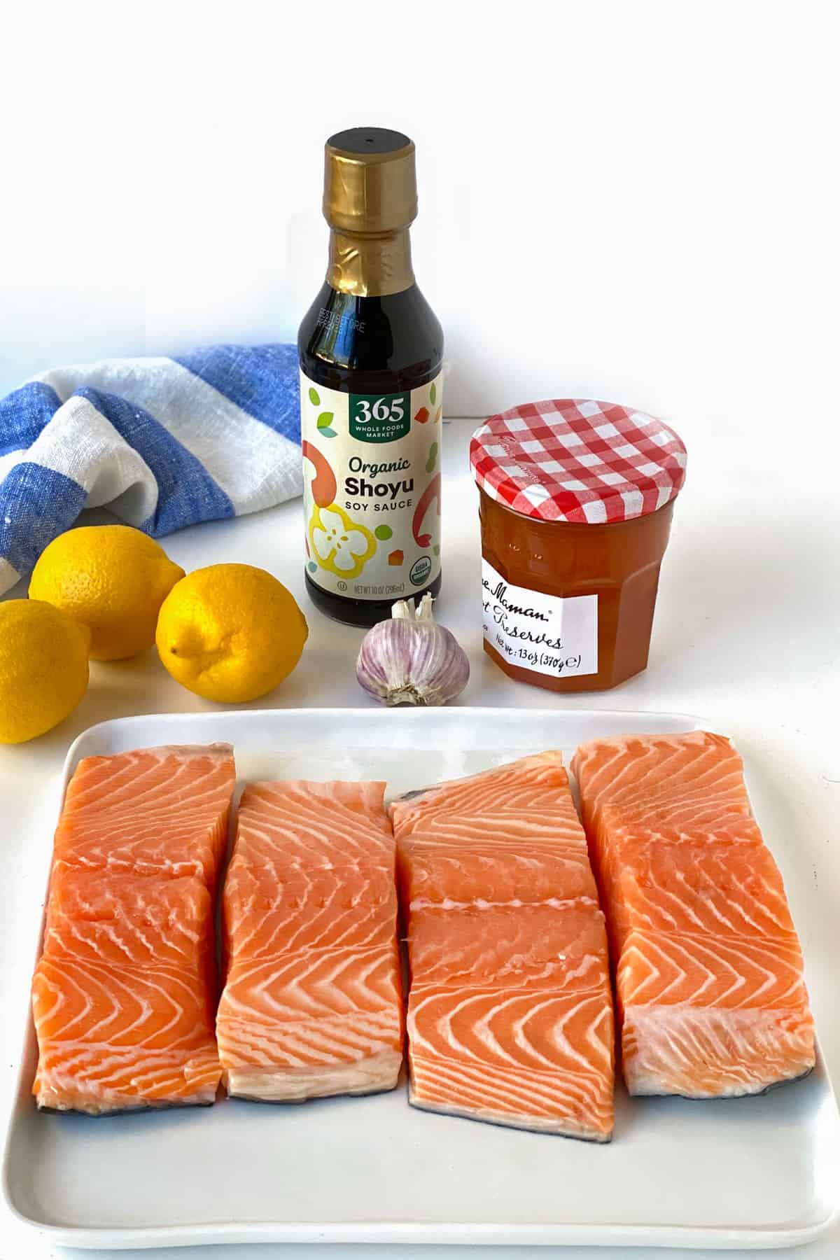 4 salmon fillets on a square white plate in the foreground, behind it are 3 whole lemons, a head of garlic, a bottle of soy sauce and a jar of apricots preserves.