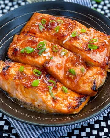 three broiled glazed salmon fillets in a black serving bowl, topped with chopped scallions