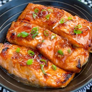 three broiled glazed salmon fillets in a black serving bowl, topped with chopped scallions