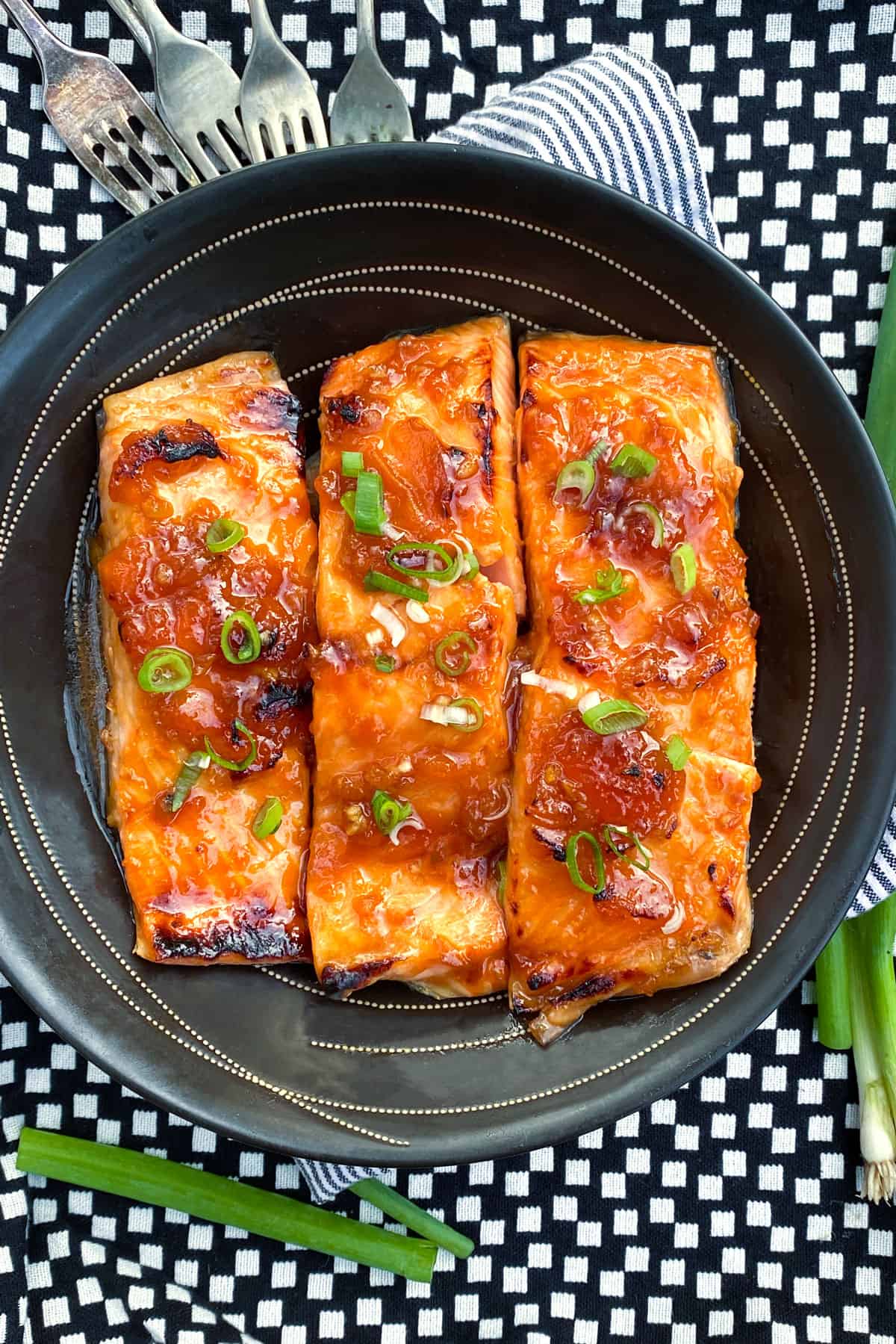 overhead shot of 3 pieces of apricot-glazed, broiled salmon in a black bowl, topped with chopped scallions, all on a vivid black and white table cloth
