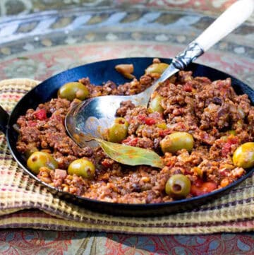 Cast iron skillet filled with ground beef stew in tomato sauce with olives and a bay leaf.