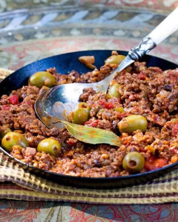 Cast iron skillet filled with ground beef stew in tomato sauce with olives and a bay leaf.