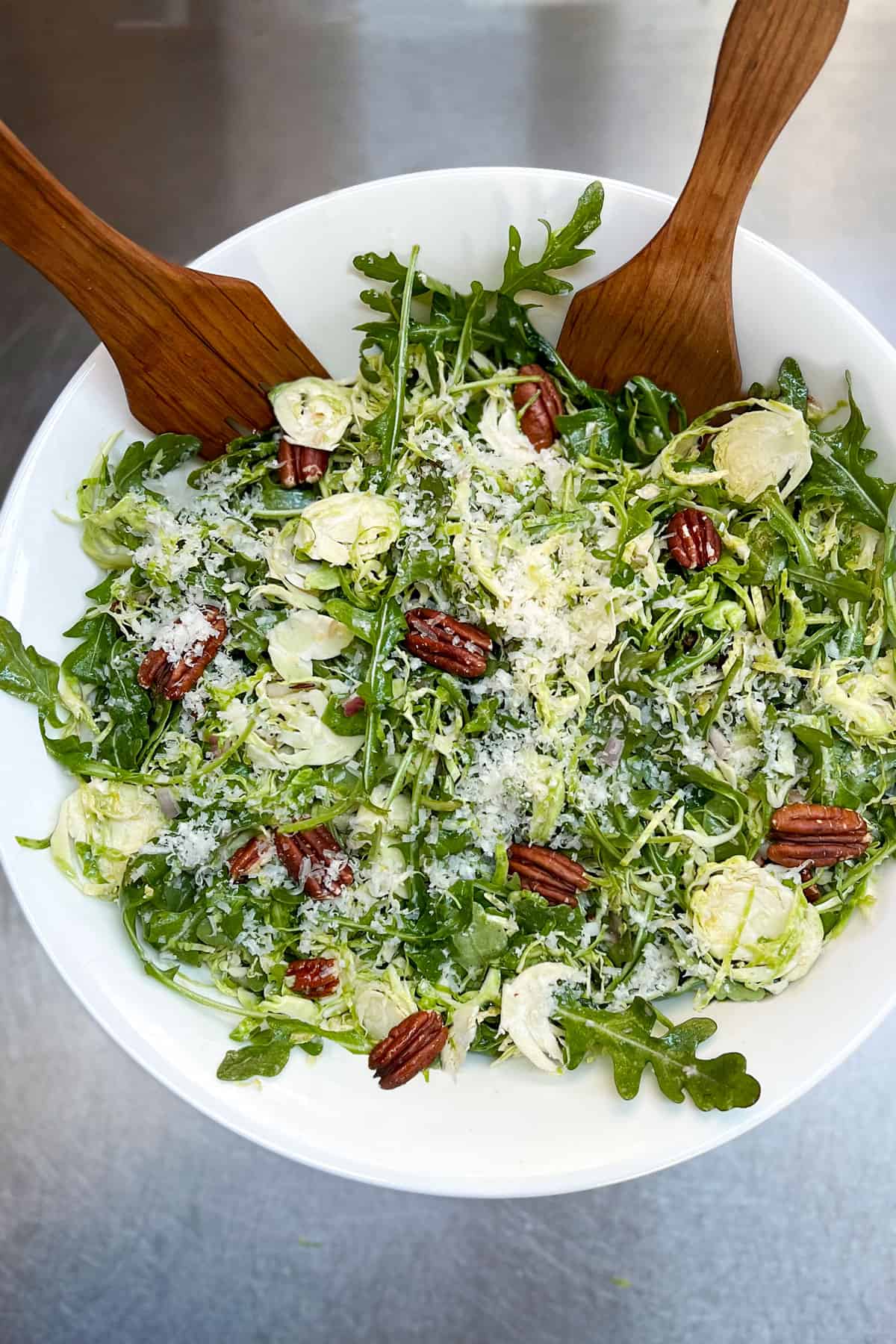 Overhead shot of a white bowl filled with shredded Brussels sprout and arugula salad with toasted pecans and grated parmesan cheese, two brown wooden salad forks stuck into the salad