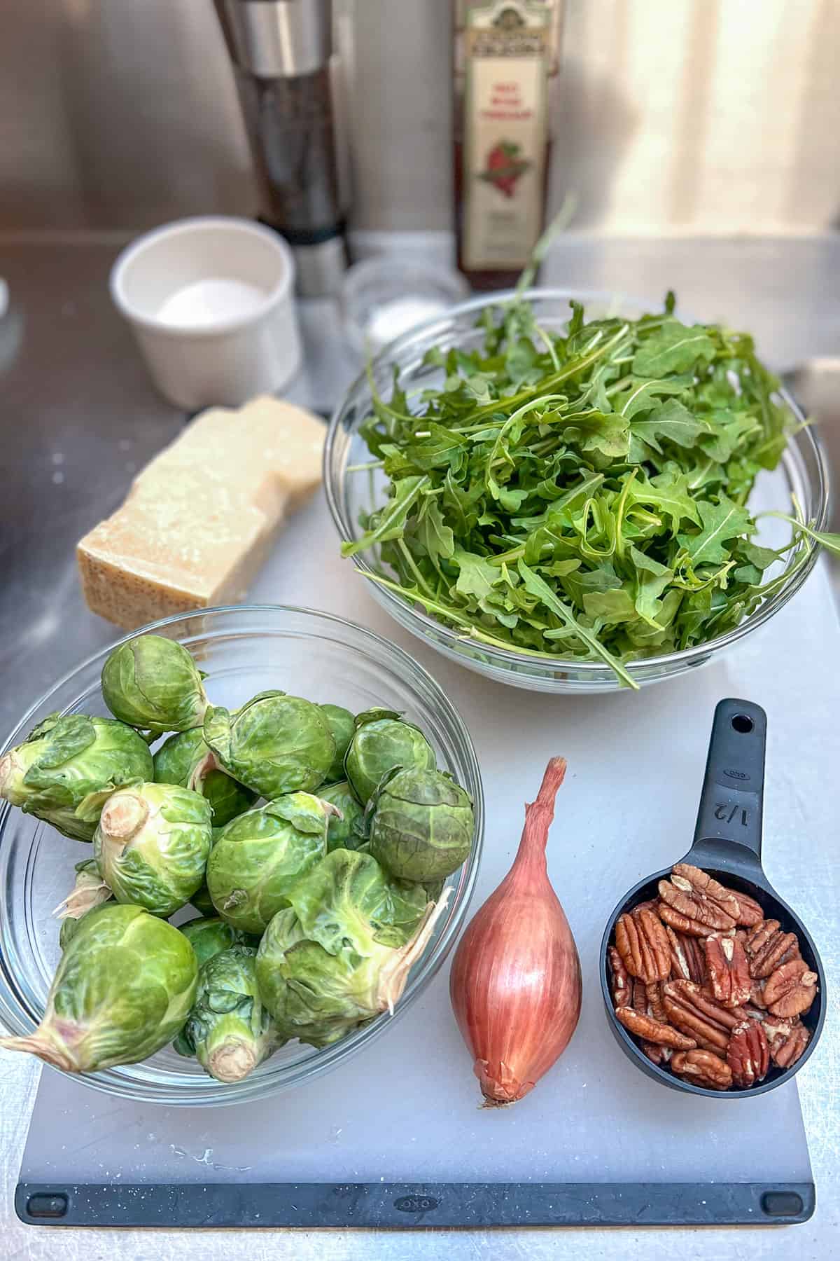 Ingredients for shaved brussels sprouts salad: white cutting board topped with a bowl of brussels sprouts, a bowl of arugula, one shallot, a small cup of pecans, a hunk of parmesan cheese 